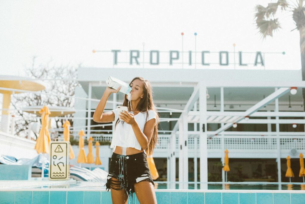 Young woman in casual wear drinking and spilling milk on body while standing near swimming pool and using mobile phone on sunny summer day